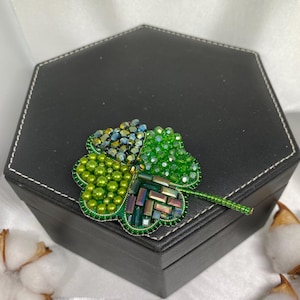 May include: A beaded four-leaf clover brooch in shades of green and iridescent accents. The brooch is made with small beads and has a detailed design. The clover is set against a black hexagonal box.
