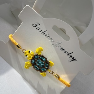 Könnte beinhalten: Ein gelbes Perlenarmband mit Schildkröte und blau-braunem Panzer. Das Armband ist an einem gelben Kordel befestigt und hat goldfarbene Verschlüsse. Die Worte "Fashion Jewelry" sind auf der weißen Karte aufgedruckt.