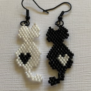 Puede incluir: Un par de pendientes de gato de cuentas negras y blancas. Los gatos se miran y tienen un diseño en forma de corazón en el pecho.