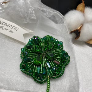 Puede incluir: Un broche de trébol de cuatro hojas hecho a mano con cuentas verdes. El broche está hecho con varias tonalidades de cuentas y cristales verdes, con un tallo de cuentas. El broche se presenta en una bolsa de organza blanca con una etiqueta que dice "NDMADE lly for You".
