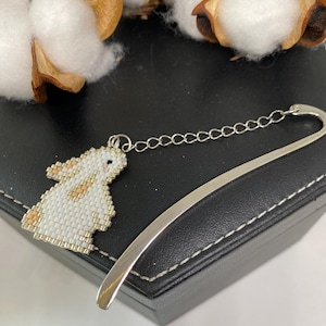 May include: A silver metal bookmark with a beaded bunny charm in white and tan. The bookmark has a curved design and a chain connecting the charm. The background includes cotton bolls and a black surface.
