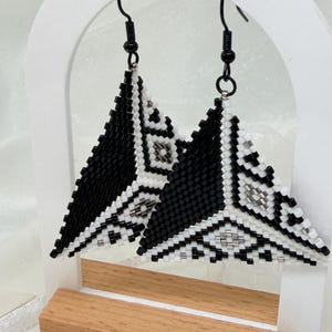 May include: A pair of triangular drop earrings crafted from small black and white beads arranged in a geometric pattern. The earrings feature black metal hooks and are suspended from a white arched display stand with a light wood base.
