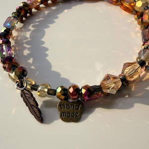 Könnte beinhalten: Ein mehrreihiges Wickelarmband mit einer Auswahl an facettierten Glasperlen in Bernstein-, Bronze- und schillernden Violetttönen. Das Armband enthält einen bronzefarbenen Metallanhänger in Federform und einen kleinen blumenförmigen Anhänger mit der Aufschrift Handmade.