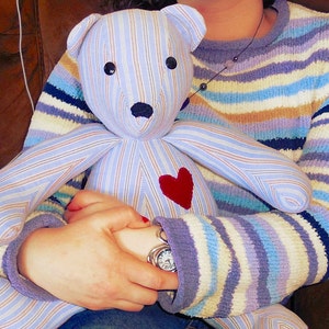 May include: A person is holding a blue and white striped teddy bear with a red heart on its chest. The teddy bear is made from fabric and has a button nose. The person is wearing a striped sweater.