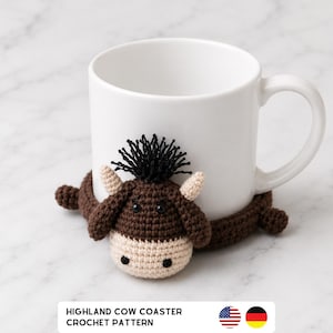 May include: A white ceramic mug rests on a handmade crochet coaster shaped like a Highland cow. The coaster features a brown body, a cream-colored face, and black fringe hair. The text "HIGHLAND COW COASTER CROCHET PATTERN" is at the bottom.