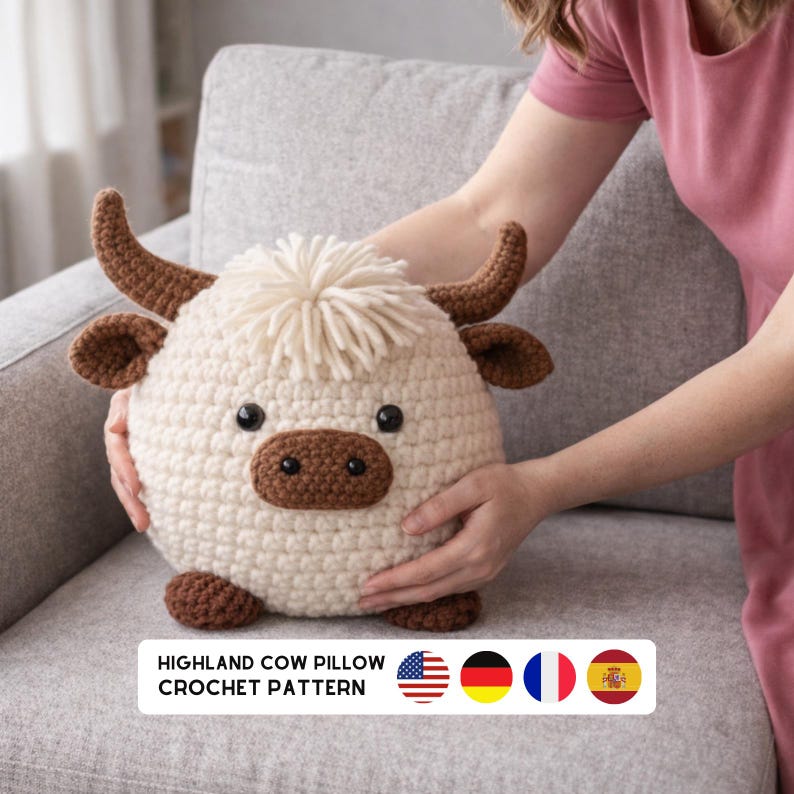 K&ouml;nnte beinhalten: Ein geh&auml;keltes Highland-Kuh-Kissen in Creme, Braun und Schwarz. Das runde Kissen hat einen flauschigen Fransen, H&ouml;rner und eine braune Schnauze. Der Text "HIGHLAND COW PILLOW CROCHET PATTERN" befindet sich unten.