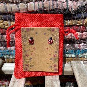Könnte beinhalten: Ein Paar rote und schwarze Marienkäfer-Ohrringe in einem roten Jutesäckchen mit einer braunen Karte, auf der "Handmade with Love" steht.