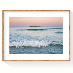 May include: A framed print of a seascape featuring a tranquil ocean scene. The image captures gentle waves breaking on the shore, with a small island visible in the distance under a pastel-colored sky. The frame is a natural wood tone.