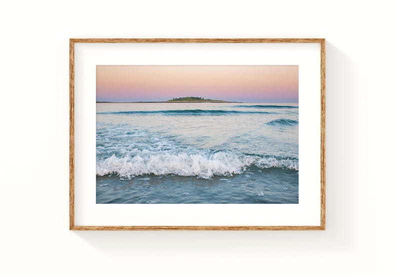 May include: A framed print of a seascape featuring a tranquil ocean scene. The image captures gentle waves breaking on the shore, with a small island visible in the distance under a pastel-colored sky. The frame is a natural wood tone.