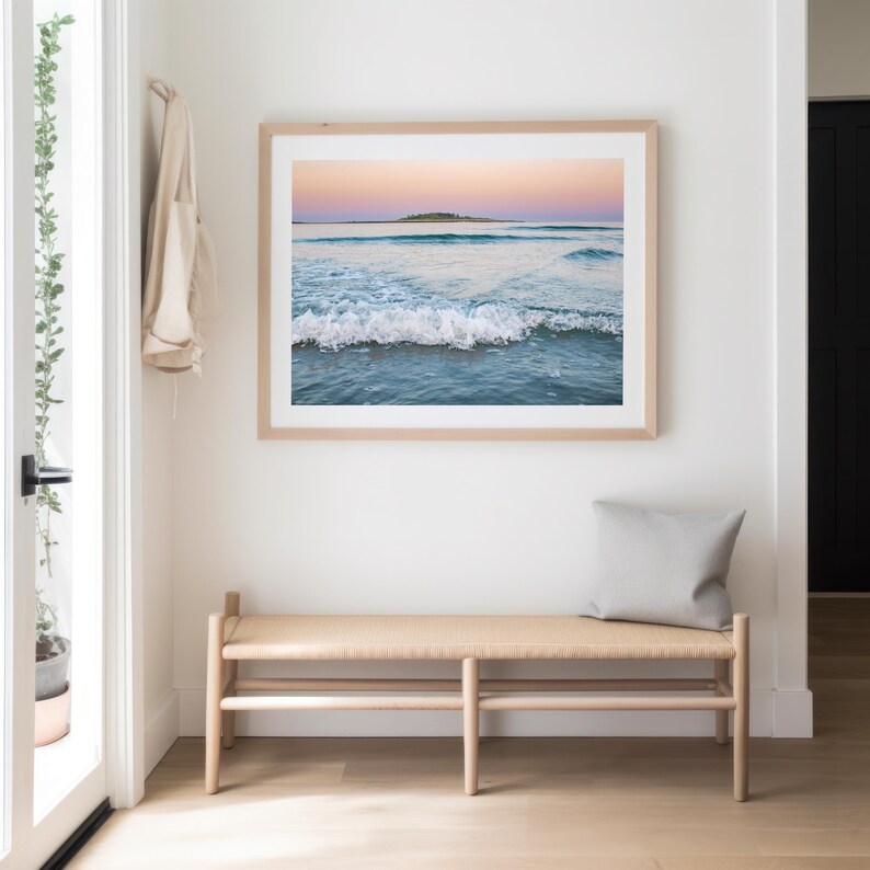 May include: A framed print of a seascape with a light wood frame hangs above a wooden bench with a gray pillow. The artwork depicts ocean waves under a pastel sky with a small island in the distance. The room has white walls and a light wood floor.