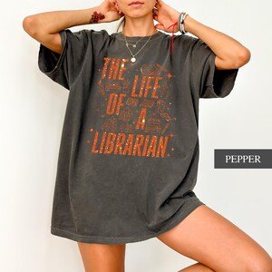 May include: Dark gray t-shirt with the text "THE LIFE OF A LIBRARIAN" in orange. The design includes illustrations of books and other library-related items. The shirt is a comfortable, oversized fit.