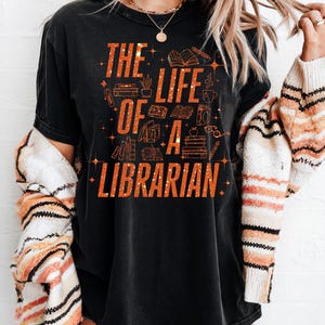 May include: Black t-shirt with the text "THE LIFE OF A LIBRARIAN" in orange glitter lettering. The design includes illustrations of books, pencils, and coffee cups. The word "BLACK" is in a black rectangle in the top left corner.