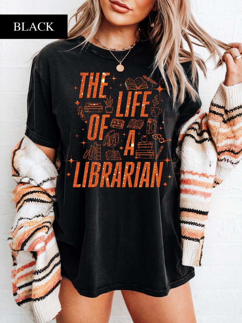 May include: Black t-shirt with the text "THE LIFE OF A LIBRARIAN" in orange glitter lettering. The design includes illustrations of books, pencils, and coffee cups. The word "BLACK" is in a black rectangle in the top left corner.