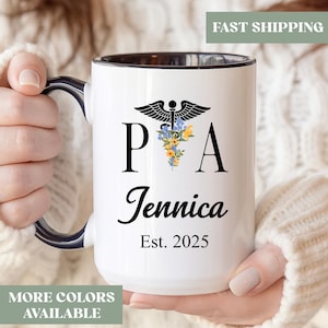 May include: White ceramic mug with a black interior and handle, featuring the letters "P A" with a floral design and the name "Jennica" and "Est. 2025" in black. The mug has a blue accent on the handle.