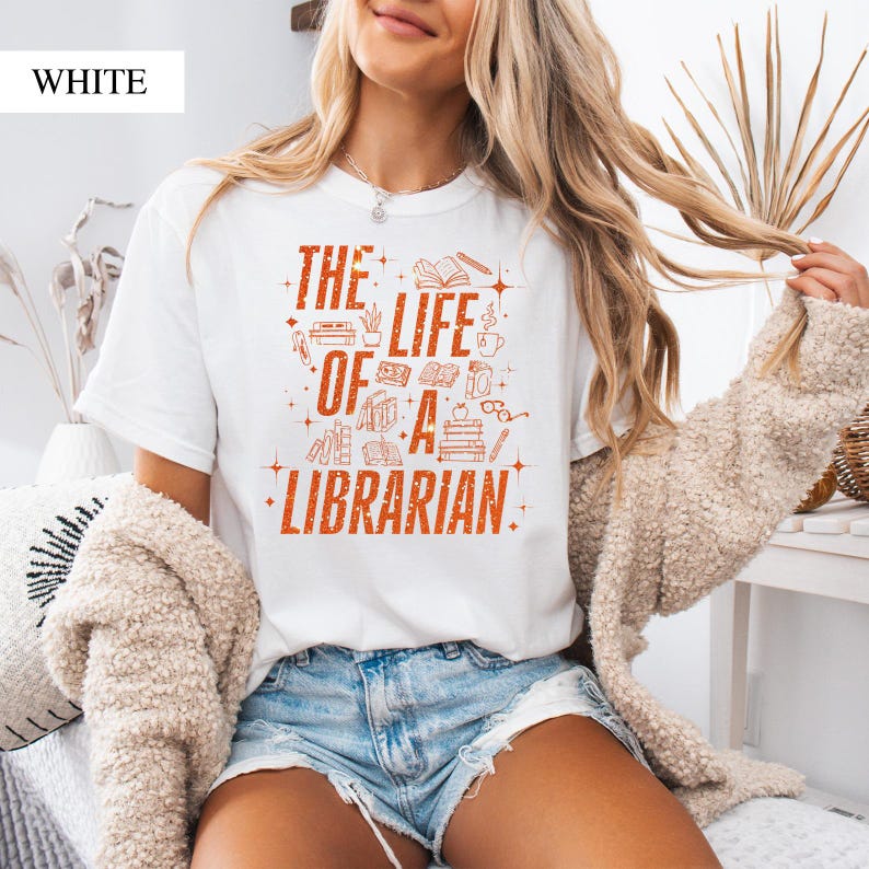 May include: White t-shirt with the text "THE LIFE OF A LIBRARIAN" in orange. The design includes illustrations of books, a coffee cup, glasses, and an apple. The shirt is a casual style.