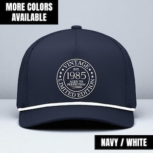 May include: Navy blue baseball cap with white accents. The cap features a white circular design with the text "VINTAGE EST. 1985 AGED TO PERFECTION LIMITED EDITION." The text "MORE COLORS AVAILABLE" is at the top, and "NAVY / WHITE" at the bottom.