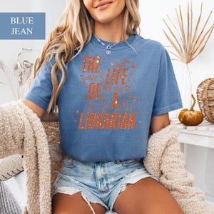 May include: A blue jean short-sleeved t-shirt with the text "THE LIFE OF A LIBRARIAN" in orange glitter lettering. The shirt has a crew neck and is made of a soft material. The shirt is a casual style.
