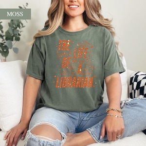 May include: A moss green t-shirt with the text "THE LIFE OF A LIBRARIAN" in orange. The shirt is a crew neck and is worn with blue jeans. The shirt is a comfortable, casual style.