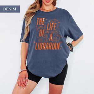 May include: Denim blue t-shirt with the text "THE LIFE OF A LIBRARIAN" in orange. The graphic includes illustrations of books and other library-related items. The shirt is a casual, oversized style.