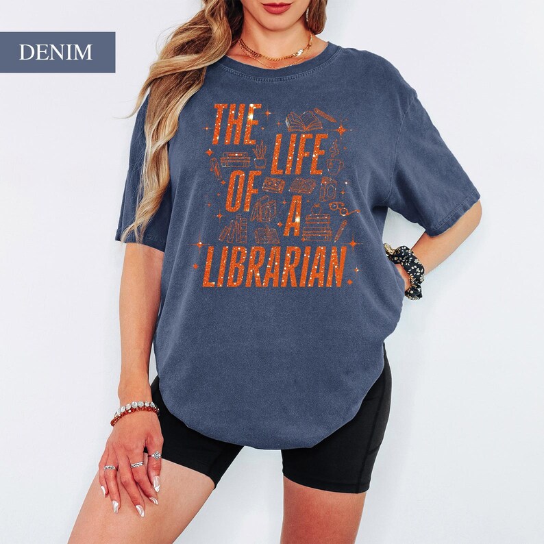 May include: Denim blue t-shirt with the text "THE LIFE OF A LIBRARIAN" in orange. The graphic includes illustrations of books and other library-related items. The shirt is a casual, oversized style.