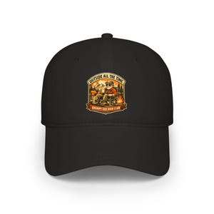 May include: A black baseball cap featuring a graphic print of a person sitting in a camping chair by a fire with mountains in the background. The text reads OUTSIDE ALL THE TIME and GRUMPY OLD MAN CLUB in a beige and orange color palette.
