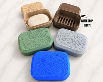 May include: Five rectangular soap dishes with textured lids in tan, brown, green, gray, and blue. One brown dish features a slotted interior and the text WITH DRIP TRAY! A tan dish contains an oval bar of soap with the embossed text Aveeno.