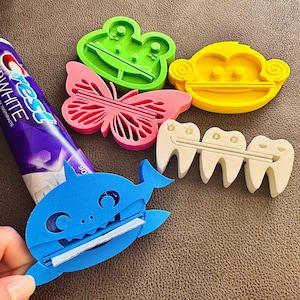 May include: A set of five colourful 3D printed toothbrush holders. The holders are shaped like a frog, a monkey, a butterfly, a set of teeth, and a blue shark. The holders are designed to hold toothbrushes and toothpaste tubes.