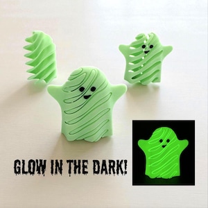 Pode incluir: Quatro figuras em forma de fantasma verde claro com olhos e sorrisos pretos. Os fantasmas têm um design texturizado e rodopiante. Uma imagem mostra o fantasma brilhando em verde no escuro. O texto na parte inferior diz "GLOW IN THE DARK!"