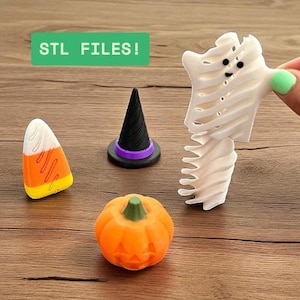 Puede incluir: Una colección de objetos impresos en 3D con temática de Halloween, que incluye un caramelo de maíz, un sombrero de bruja, una calabaza tallada y un fantasma. Los objetos son de varios colores y están sobre una superficie de madera. El texto "STL FILES!" está en un rectángulo verde.