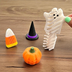 Puede incluir: Una colección de decoraciones temáticas de Halloween, que incluyen un caramelo de maíz, un sombrero de bruja con una banda morada, una calabaza naranja con una cara tallada y una figura blanca en forma de fantasma. Los artículos están dispuestos sobre una superficie de madera.