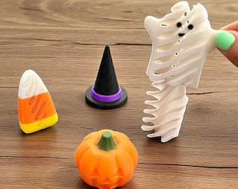 May include: A collection of Halloween-themed decorations, including a candy corn, a witch's hat with a purple band, an orange pumpkin with a carved face, and a white ghost-shaped figure. The items are arranged on a wooden surface.