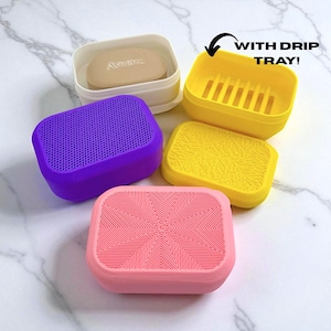 May include: A collection of colorful soap dishes in various shades including pink, purple, yellow, and white. One dish contains a bar of soap. The yellow dish has a drip tray. The text "WITH DRIP TRAY!" is visible.