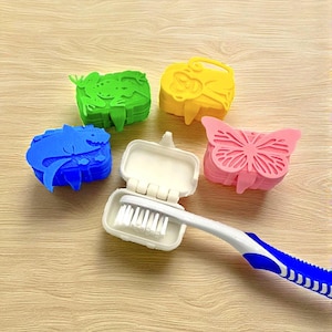 May include: A toothbrush with a white and blue handle rests in an open white travel case. Four colorful, animal-shaped toothbrush holders in blue, green, yellow, and pink surround the case. The holders are shaped like a shark, frog, monkey, and butterfly.