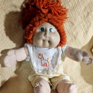 May include: A Cabbage Patch Kid doll with red yarn hair, wearing a yellow and white outfit with a giraffe embroidered on the bib. The doll has green eyes and a rosy complexion.