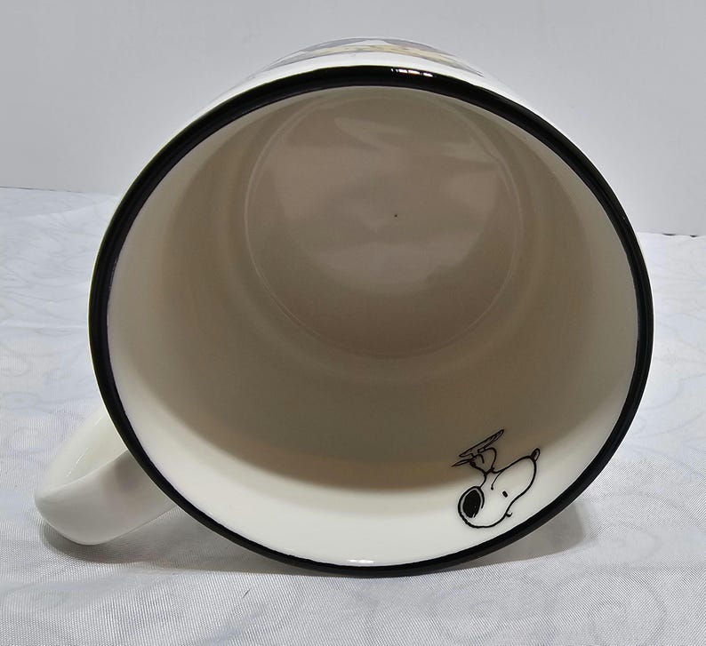 Pu&ograve; includere: Tazza in ceramica bianca con bordo e manico neri. L'interno presenta un'illustrazione a cartoni animati di Snoopy con cappello e occhiali da aviatore. La tazza &egrave; progettata per bevande calde e ha un design classico.