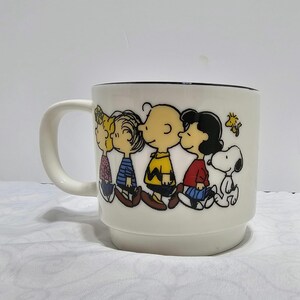 Pu&ograve; includere: Tazza in ceramica bianca con un'illustrazione a cartoni animati dei personaggi di Peanuts: Charlie Brown, Lucy, Linus e Snoopy. La tazza ha un bordo nero e un manico bianco. I personaggi indossano i loro abiti classici.