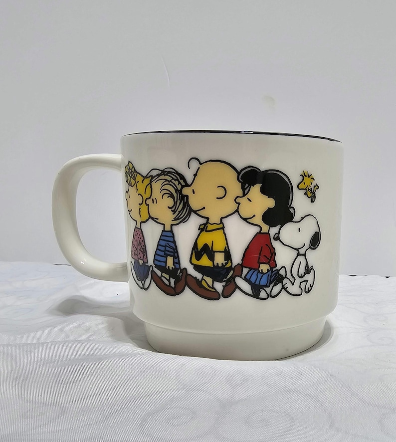 Pu&ograve; includere: Tazza in ceramica bianca con un'illustrazione a cartoni animati dei personaggi di Peanuts: Charlie Brown, Lucy, Linus e Snoopy. La tazza ha un bordo nero e un manico bianco. I personaggi indossano i loro abiti classici.