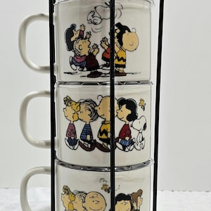 May include: A set of three white ceramic mugs featuring Peanuts characters, stacked in a black metal stand. Each mug has a different illustration of Charlie Brown, Snoopy, Lucy, and other characters. The mugs have a curved handle.