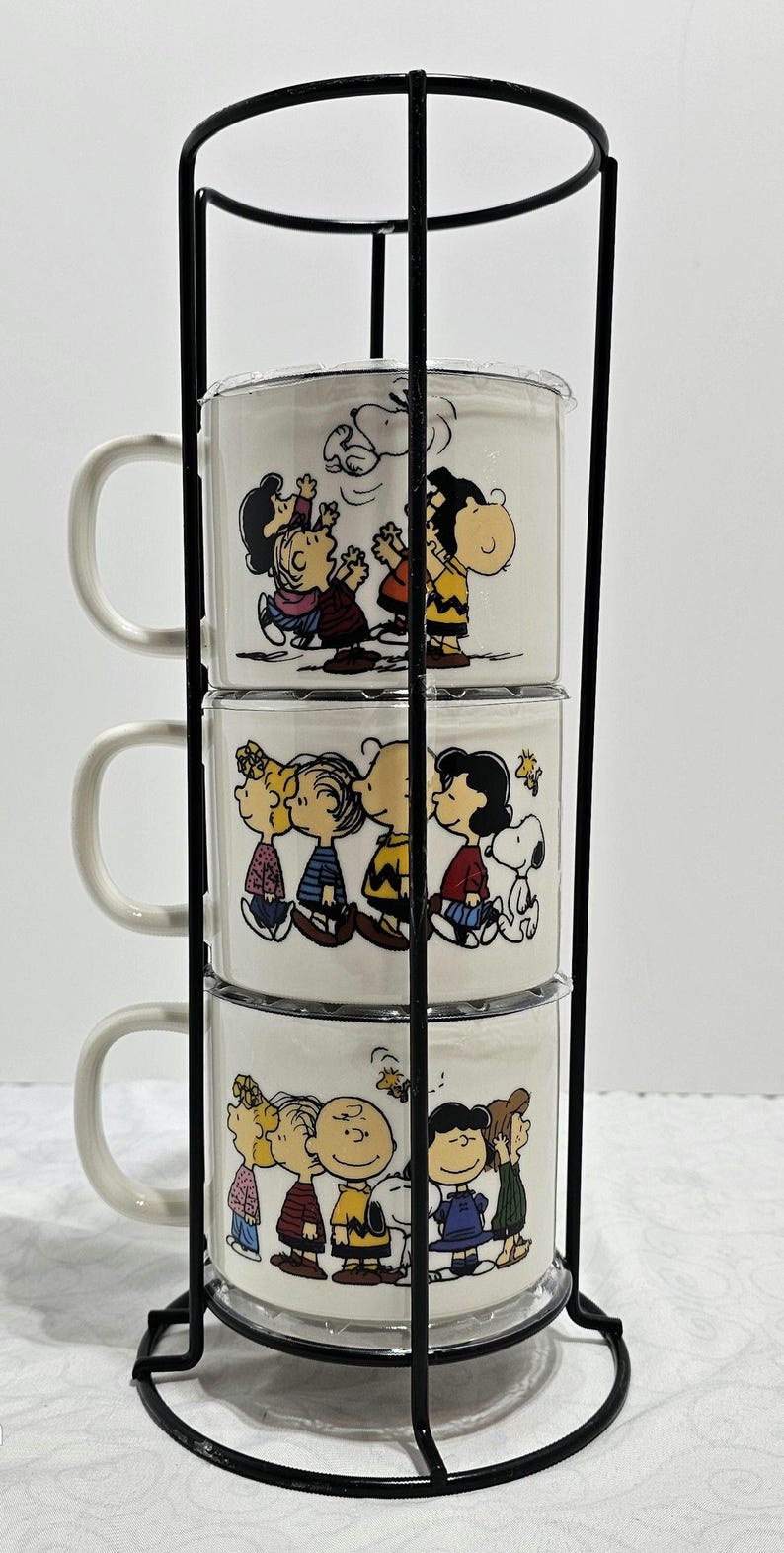 Pu&ograve; includere: Un set di tre tazze in ceramica bianca con personaggi dei Peanuts, impilate in un supporto in metallo nero. Ogni tazza presenta un'illustrazione diversa di Charlie Brown, Snoopy, Lucy e altri personaggi. Le tazze hanno un manico curvo.