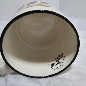 Pu&ograve; includere: Tazza in ceramica bianca con bordo e manico neri. La tazza presenta un'illustrazione a cartoni animati di Snoopy e Woodstock. L'interno &egrave; bianco e la tazza &egrave; angolata verso l'osservatore.