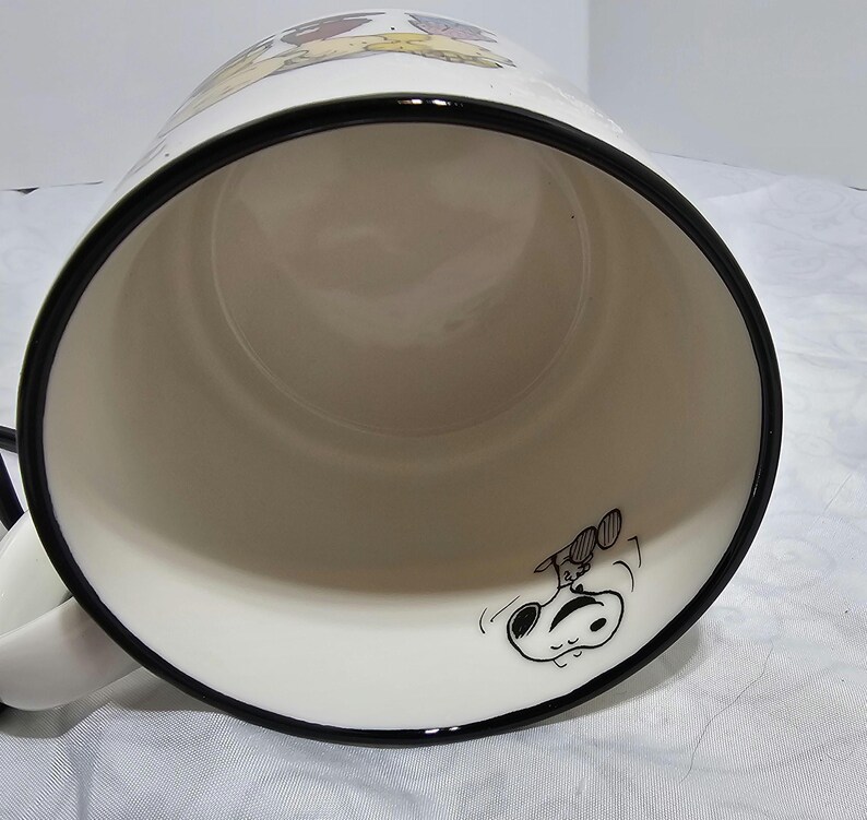 Pu&ograve; includere: Tazza in ceramica bianca con bordo e manico neri. La tazza presenta un'illustrazione a cartoni animati di Snoopy e Woodstock. L'interno &egrave; bianco e la tazza &egrave; angolata verso l'osservatore.