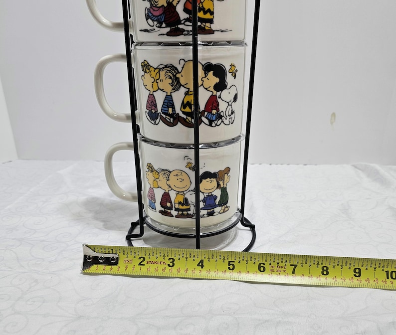 Pu&ograve; includere: Una pila di tre tazze di ceramica bianca con personaggi dei cartoni animati, tenute in un supporto di metallo nero. Ogni tazza mostra un diverso gruppo di personaggi con abiti colorati. Le tazze sono impilate verticalmente.
