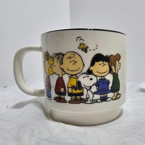 Pu&ograve; includere: Una tazza in ceramica bianca con bordo nero e manico. La tazza presenta un'illustrazione colorata della banda dei Peanuts, tra cui Charlie Brown, Snoopy e Lucy. La tazza &egrave; di dimensioni standard.