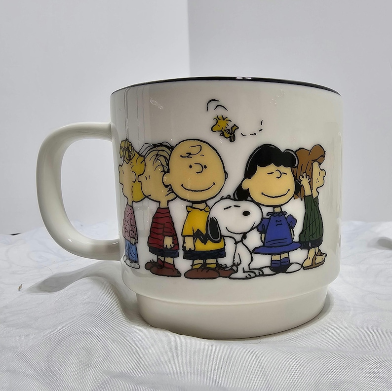 Pu&ograve; includere: Una tazza in ceramica bianca con bordo nero e manico. La tazza presenta un'illustrazione colorata della banda dei Peanuts, tra cui Charlie Brown, Snoopy e Lucy. La tazza &egrave; di dimensioni standard.