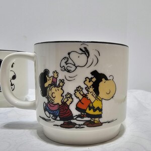 Pu&ograve; includere: Tazza in ceramica bianca con bordo nero, raffigurante un'illustrazione colorata dei personaggi di Peanuts. Snoopy &egrave; mostrato a mezz'aria, con Charlie Brown e gli amici che guardano in alto. La tazza ha un manico e una base larga.