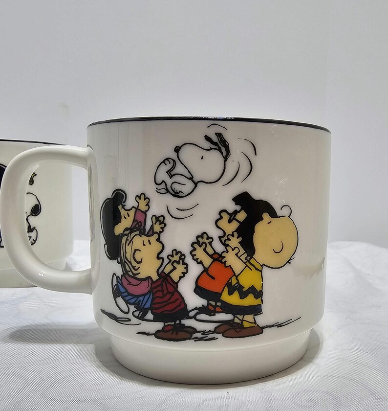 Pu&ograve; includere: Tazza in ceramica bianca con bordo nero, raffigurante un'illustrazione colorata dei personaggi di Peanuts. Snoopy &egrave; mostrato a mezz'aria, con Charlie Brown e gli amici che guardano in alto. La tazza ha un manico e una base larga.
