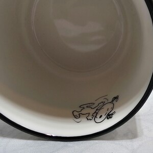 Pu&ograve; includere: Una ciotola di ceramica bianca con bordo nero. Un'illustrazione a cartoni animati in bianco e nero di Snoopy &egrave; sul lato della ciotola. La ciotola &egrave; vuota e sembra pulita.