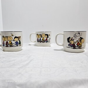 Pu&ograve; includere: Tre tazze in ceramica bianca con bordi e manici neri. Ogni tazza presenta un'illustrazione diversa dei personaggi di Peanuts. Le tazze sono appoggiate su una superficie bianca.