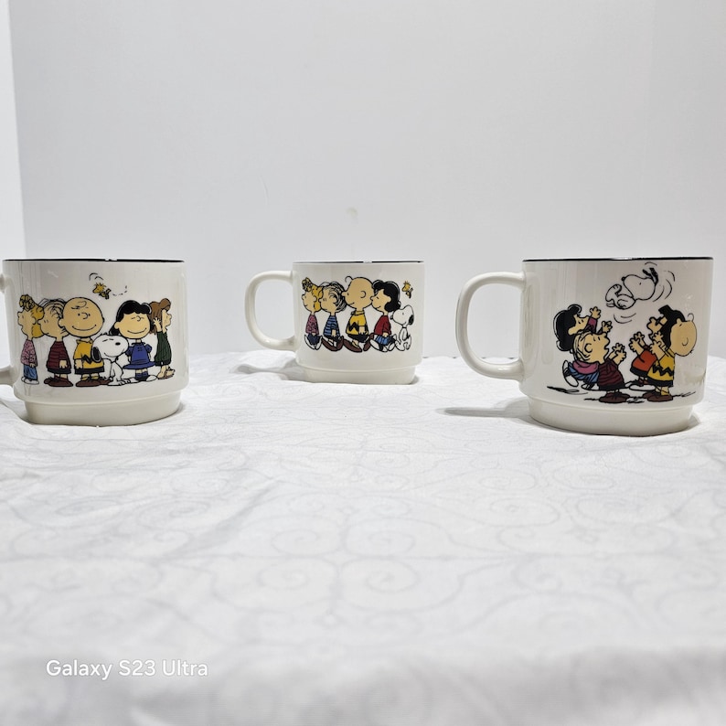 Pu&ograve; includere: Tre tazze in ceramica bianca con bordi e manici neri. Ogni tazza presenta un'illustrazione diversa dei personaggi di Peanuts. Le tazze sono appoggiate su una superficie bianca.