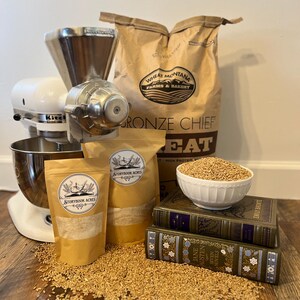 May include: A still-life image featuring baking ingredients and equipment. A white KitchenAid mixer with a grain grinder attachment is next to bags of wheat and Storybook Acres flour. A bowl of wheat berries sits atop two antique books. Wheat kernels are scattered on the wooden surface.